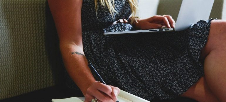 A woman holding a laptop and writing in a notebook.