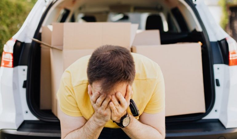 A man with his head in his hands, with moving boxes in the trunk of his car.
