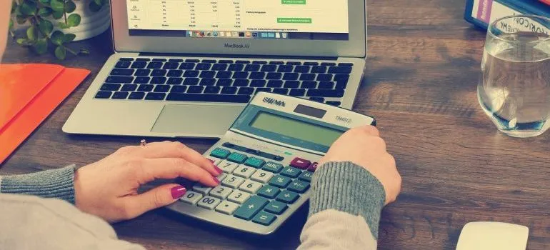 A woman typing on a calculator in front of a laptop, preventing one of moving mistakes for newlyweds.
