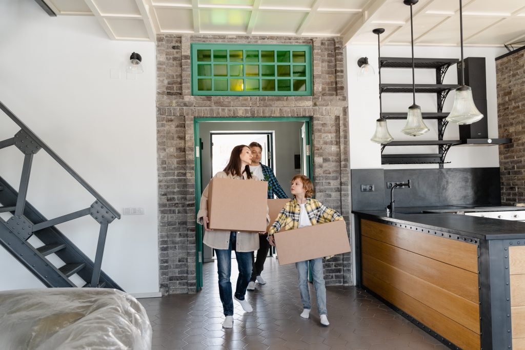 family of three carrying moving boxes, entering a house