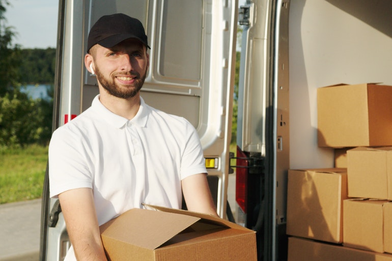 A man in a white shirt carrying a box in front of a back of a white moving van.
