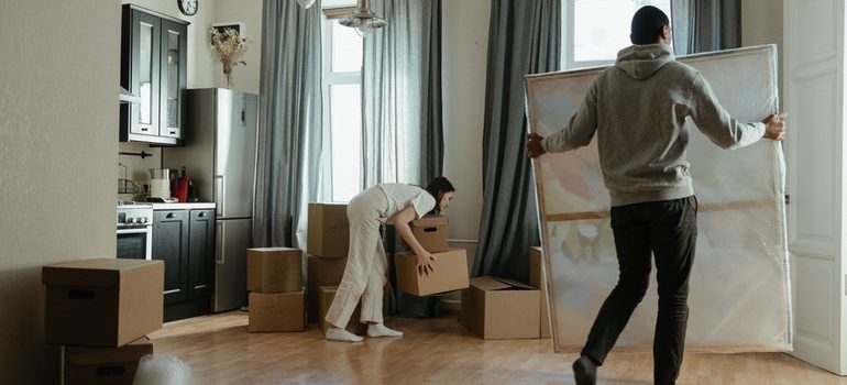 A man carrying a packed painting and a woman bent over picking up a box in a half-packed apartment.