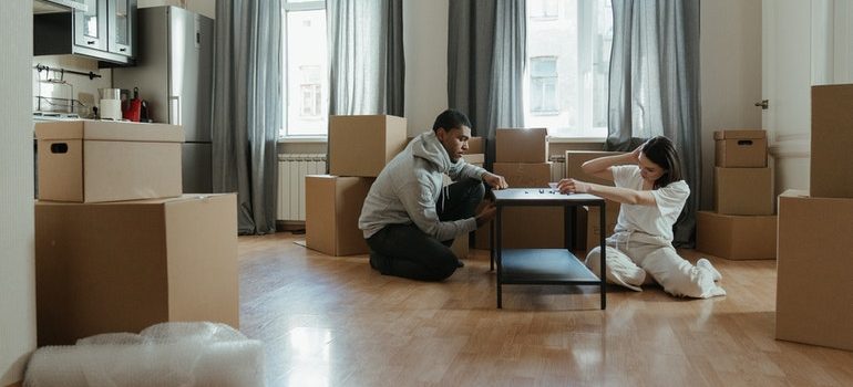 A man and a woman sitting on a floor by a table in a half packed apartment.