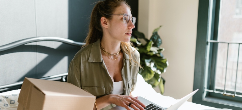 a gilrl sitting on th ebed with her laptop thinking of ways to speed up the unpacking process