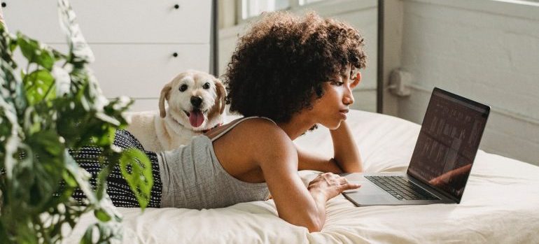 A woman looking at her laptop, dog next to her on the bed