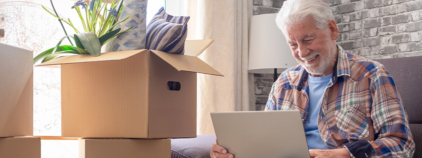 An older man sitting on a couch, focused on his laptop, with a cozy living room setting in the background.