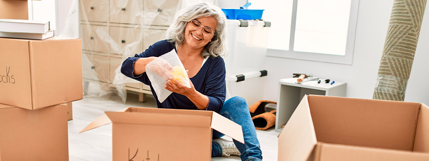 A woman sits on the floor surrounded by boxes, preparing for a move, with a focus on the challenges of relocating seniors.
