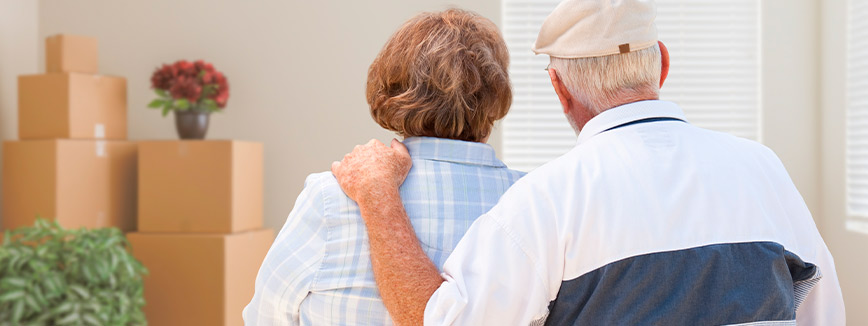 A man and woman stand together in front of moving boxes, preparing for a senior relocation.