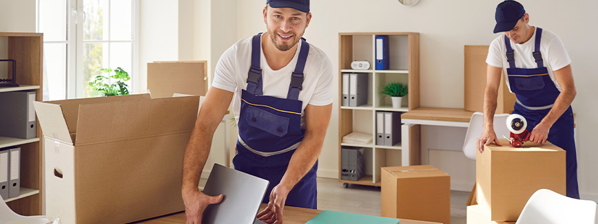 Two men in blue overalls are engaged in moving boxes, highlighting the logistics of relocating a business.
